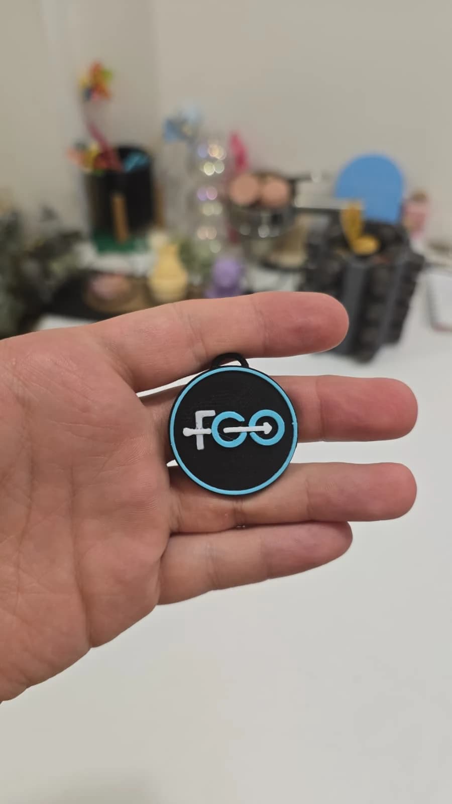 Brelocul FGO real, ținut în palmă: disc negru cu contur turcoaz, siglă albă și turcoaz și buclă neagră.